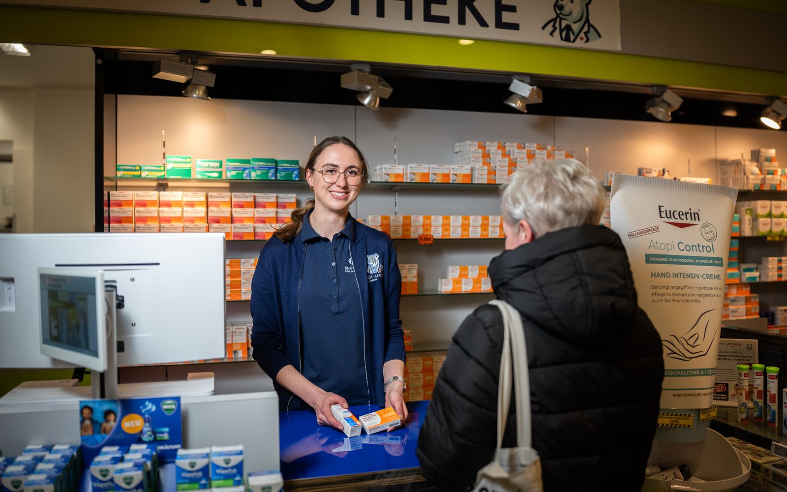 Mitarbeiterin der Knut-Apotheke lächelt am Handverkaufstisch und zeigt einer Kundin ein Produkt